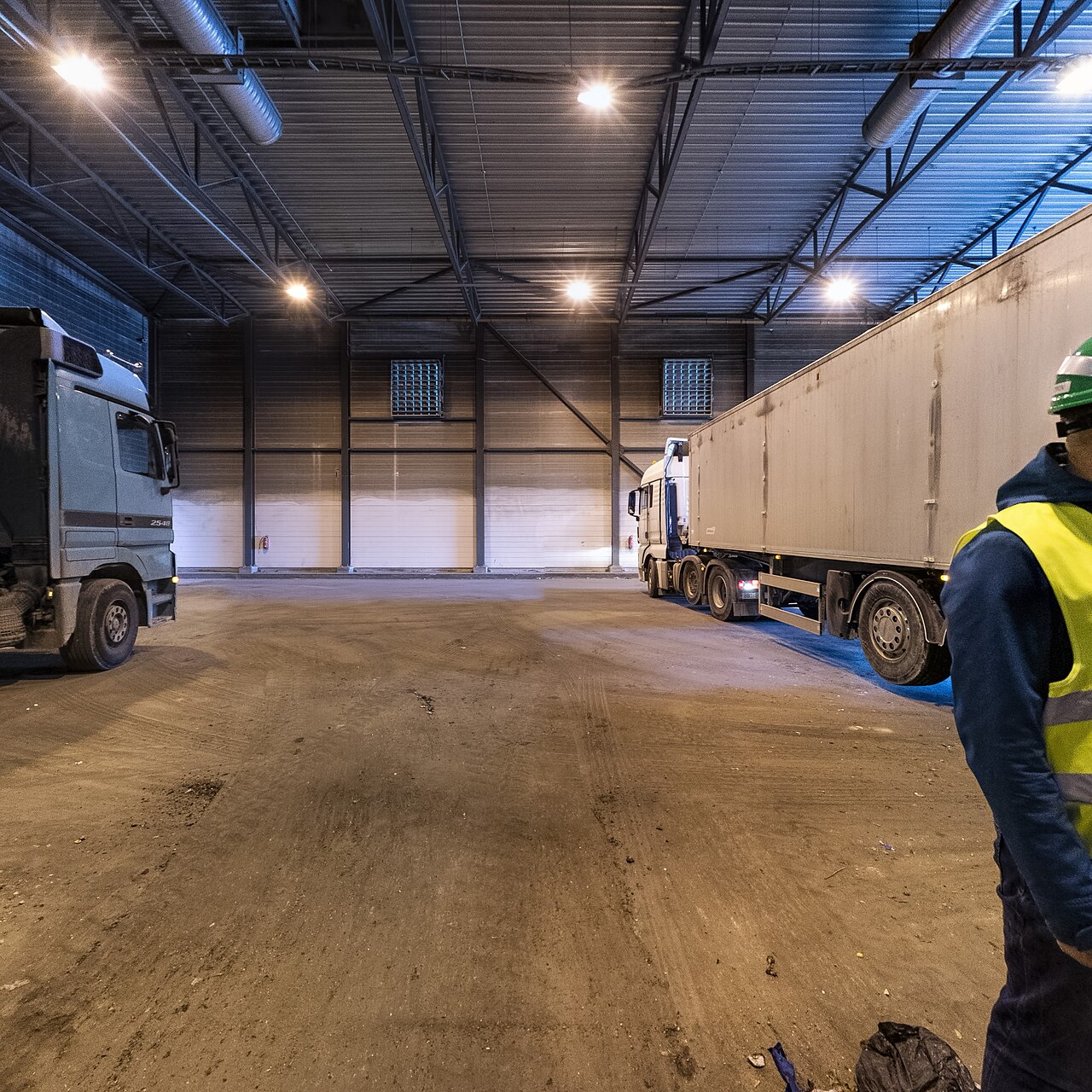 Lorries inside the receiving hall of a waste recovery plant, with a hard-hatted worker in the foreground